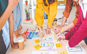 Group of designers brainstorming and sketching wireframes with sticky notes, color samples, and prototypes, representing the creative process of building custom websites and apps.
